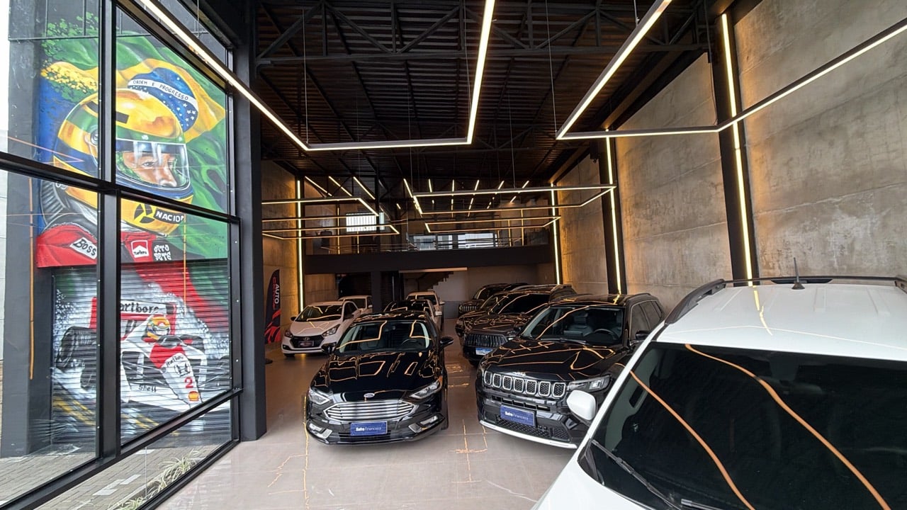 Salão ocupado por veículos, com iluminação geométrica e mezanino ao fundo